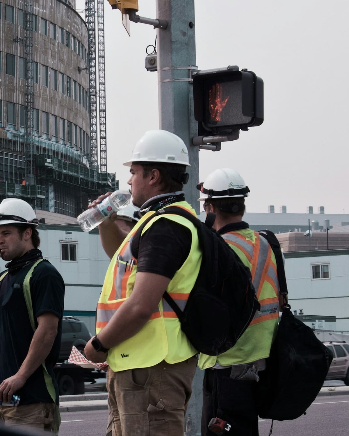 Three workers in white hard hats and hi-vis vests taking a water break at a street corner by a job site