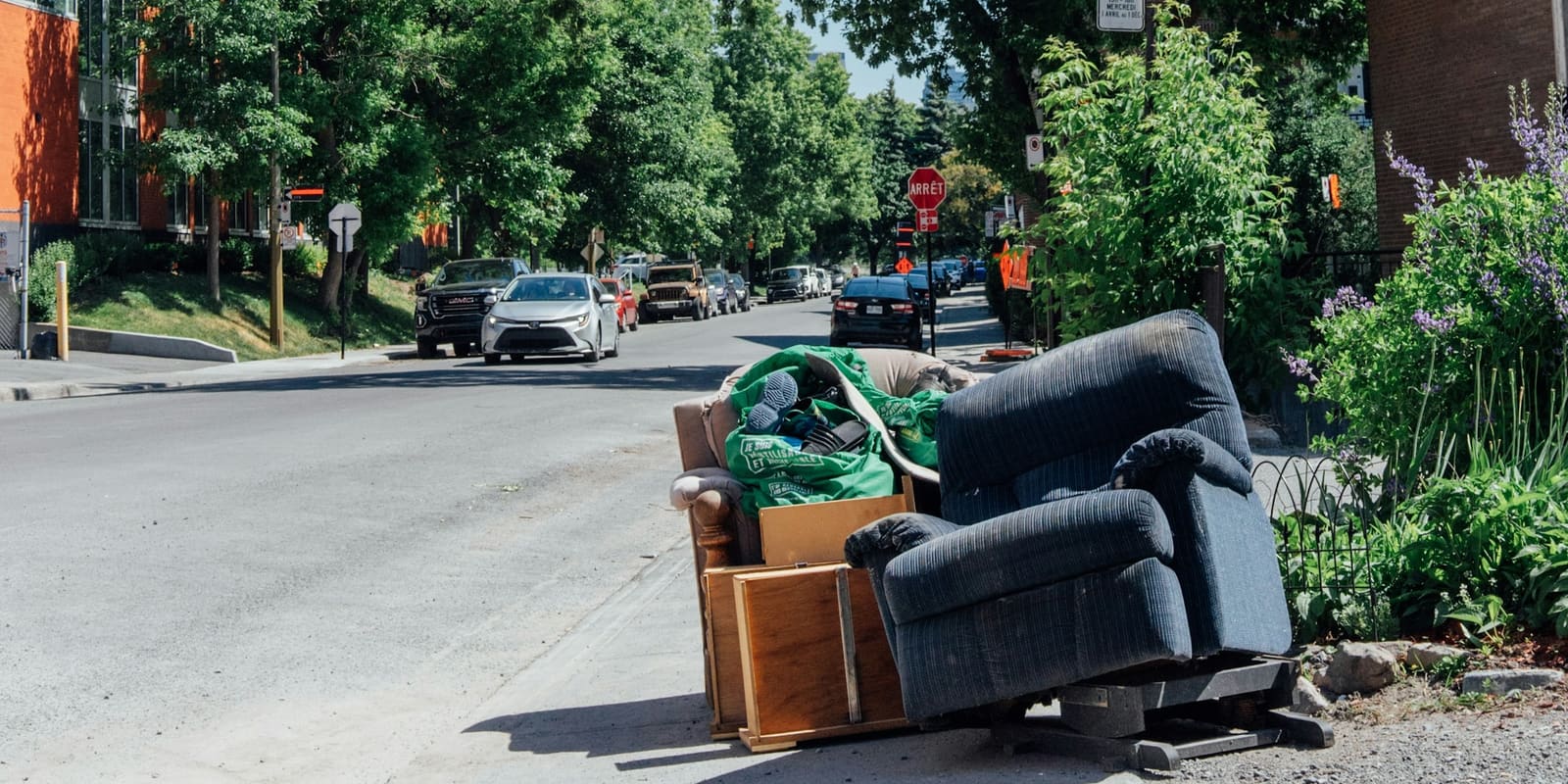 Old recliner, couch, drawers and bags of junk piled at the curb on a tree-lined residential street